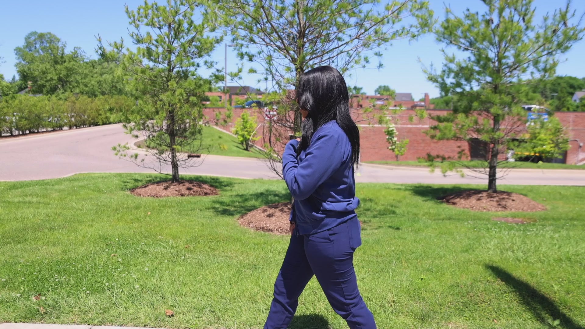 Load video: a female nurse walking outside the hospital campus. In navy blue scrubs, with a NurseBands Signature scrub jacket with embroidered logo on front and back. The grass is green the wind is blowing softly. Shes happily walking in the neighborhood. Nurse outfit, scrub set